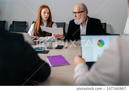 Portrait of mature adult male boss team leader giving paper report business plan to employees group at corporate briefing table. Corporate meeting office workers in modern coworking space. 107168967