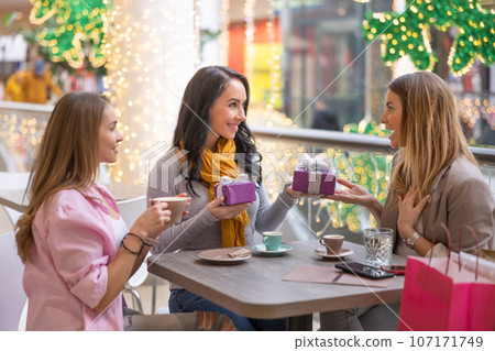 Three good-looking women are sitting in a cafe in a shopping mall, handing out Christmas presents 107171749