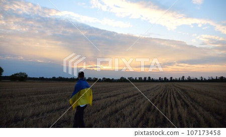 Ukrainian man walking on barley meadow with a blue-yellow banner on shoulders at sunset. Young guy going on wheat field with a flag of Ukraine as symbol of victory against russian aggresion. Slow mo 107173458