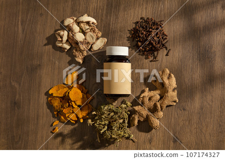On the vintage wooden background, a unlabeled bottle displayed and surrounded by folk medicines: ginger, sand ginger, cloves. Advertising scene for product, traditional medicine to supplement health 107174327