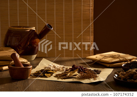 Concept of dividing herbs according to medicinal scale for daily intake. A medicinal kettle, a wooden mortar and pestle, and a sheet of paper containing various traditional medicines decorated on wood Concept of dividing herbs according to medicinal scale for daily intake. A medicinal kettle, a wooden mortar and pestle, and a sheet of paper containing various traditional medicines decorated on wood 107174352