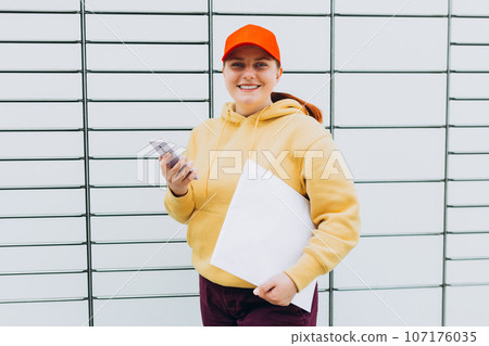 Young woman in red hat picks up parcel from automatic post office machine, Courier standing with phone and small box. Concept of fast delivery to automatic self lockers 107176035