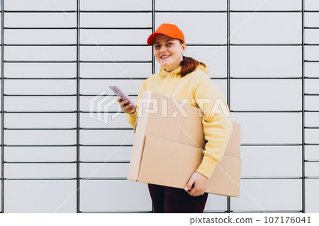 Young woman in red hat picks up parcel from automatic post office machine, Courier standing with phone and small box. Concept of fast delivery to automatic self lockers 107176041