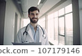 Hospital. Health care. Portrait of young smiling 30-year-old bearded caucasian man doctor wearing white medical coat with stethoscope around his neck, looking at camera. Hospital Corridor, AI 107179146
