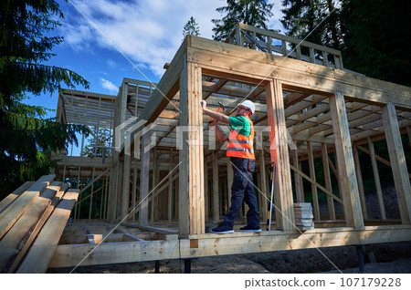 Carpenter constructing two-story wooden frame house near the forest. Man hammering nails into structure while wearing protective helmet and construction vest. Concept of modern ecological construction 107179228