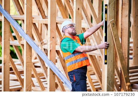 Carpenter constructing wooden frame house. Man inspects walls for levelness using spirit level, wearing protective overalls, helmet and orange vest. Concept of eco-friendly contemporary construction. Carpenter constructing wooden frame house. Man inspects walls for levelness using spirit level, wearing protective overalls, helmet and orange vest. Concept of eco-friendly contemporary construction. 107179230