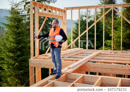 Manager overseeing construction of wooden two-story house located close to woods. Bearded man in glasses studying construction plan. Objective is to implement modern ecological construction methods. Manager overseeing construction of wooden two-story house located close to woods. Bearded man in glasses studying construction plan. Objective is to implement modern ecological construction methods. 107179231