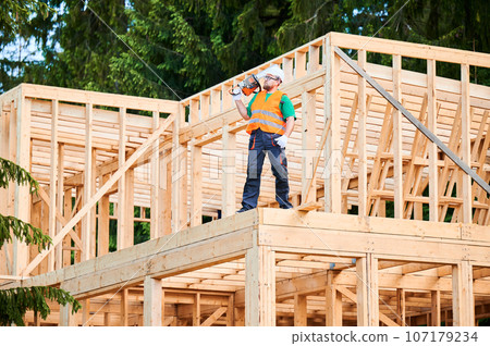 Carpenter building wooden frame two-story house. Bearded man wearing protective goggles carrying chainsaw on his shoulder, dressed in protective helmet, coveralls, and orange vest. 107179234