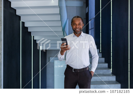 Mature experienced african american man inside office at workplace walking around office with phone in hand, portrait of businessman in shirt, smiling and looking at camera using app on smartphone. 107182185