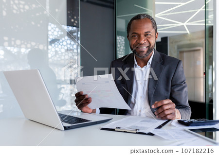 Portrait of successful mature experienced financier, business man behind paper work smiling and looking at camera, african american man at workplace reviewing contracts, accounts and reports. 107182216