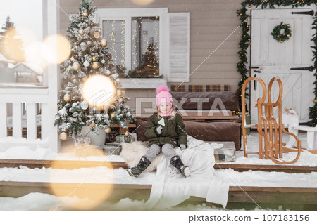 A little girl in winter clothes is sitting on the porch of a house decorated for the Christmas holidays. Christmas holidays for a child. A little girl in winter clothes is sitting on the porch of a house decorated for the Christmas holidays. Christmas holidays for a child. 107183156