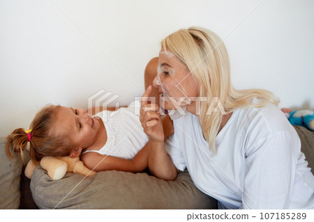 A young mother with a fabric mask on her face communicates with her daughter on the sofa at home. 107185289
