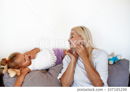 A young mother with a fabric mask on her face communicates with her daughter on the sofa at home. 107185290