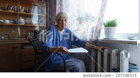 The senior woman holding gas bill in front of heating radiator. Payment for heating in winter. 107186315