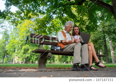The grandfather and granddaughter are looking at photos on notebook, sitting on a bench in a city park. 107187151