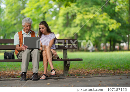 The grandfather and granddaughter are looking at photos on a notebook, sitting on a bench in a city park. 107187153