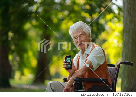 Senior diabetic man in wheelchair checking his glucose data on smartphone outdoors, in park. 107187163