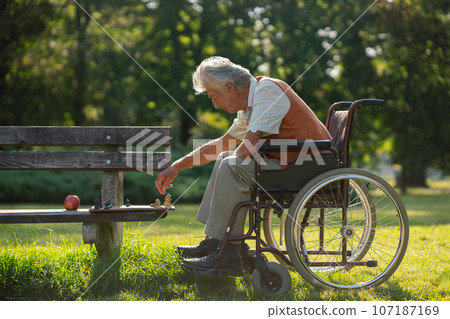 A senior man in a wheelchair with chess on bench is alone in a city park, feeling lonely. A senior man in a wheelchair with chess on bench is alone in a city park, feeling lonely. 107187169