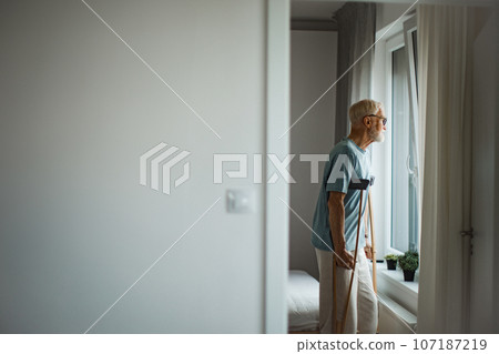 Senior man with crutches spending time alone in his apartment. Senior man with crutches spending time alone in his apartment. 107187219