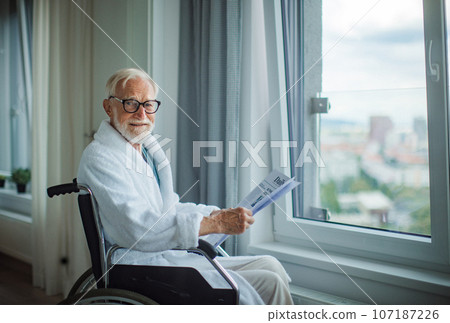 Elderly man in a wheelchair reading the newspaper in his robe in the morning. Elderly man in a wheelchair reading the newspaper in his robe in the morning. 107187226