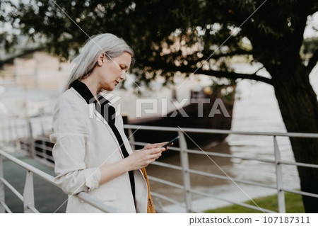 Portrait of beautiful woman with gray hair in city, using her smartphone. Portrait of beautiful woman with gray hair in city, using her smartphone. 107187311