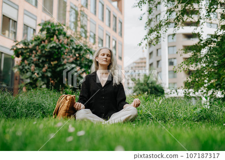 Portrait of beautiful smiling woman with gray hair meditating on the grass in city park. 107187317