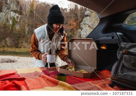 happy woman eating pizza drinking tea with view of autumn carpathian river 107187331