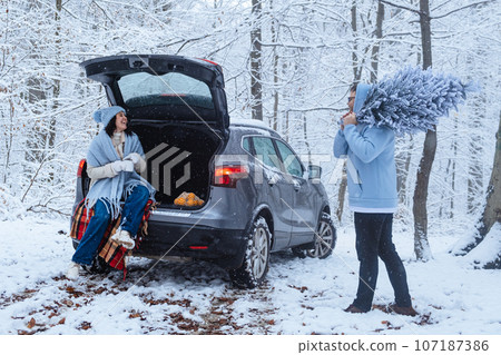 a woman sits in the trunk of a car in a winter forest, a man carries a Christmas tree on his shoulders 107187386