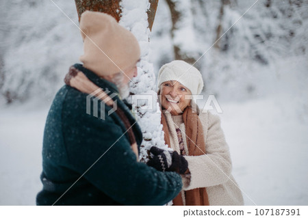 Elegant senior couple standing by a tree in the snowy park, during cold winter snowy day. 107187391