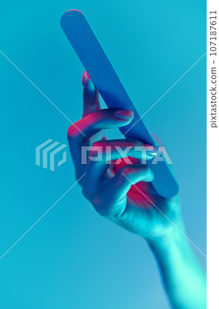 Female hand with nude, natural nails, manicure holding nail file isolated over blue background in neon light 107187611