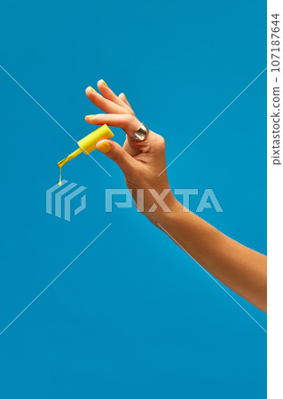 Tender female hand holding yellow nail polish with color drop against blue background. Bright manicure 107187644