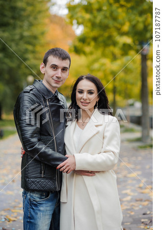A man hugs his pregnant wife. A man in a black leather jacket hugs and kisses a woman in a white coat. Autumn A man hugs his pregnant wife. A man in a black leather jacket hugs and kisses a woman in a white coat. Autumn 107187787