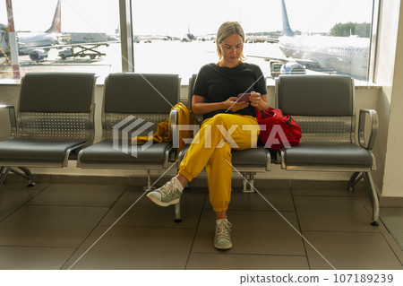 Airport terminal. Woman waiting for flight using smartphone. Girl with cell phone in airport surfing internet social media apps. Traveling female in boarding lounge of airline hub. Traveling girl Airport terminal. Woman waiting for flight using smartphone. Girl with cell phone in airport surfing internet social media apps. Traveling female in boarding lounge of airline hub. Traveling girl 107189239