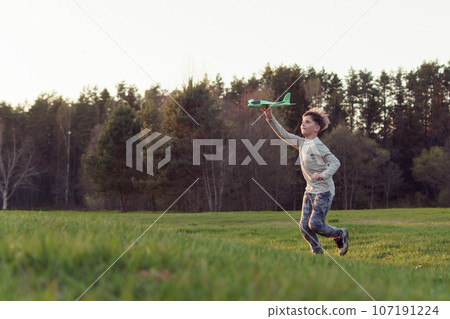 Gorgeous teenage boy running across meadow, keeping arm up and playing with toy plane, enjoying adventure in nature. 107191224