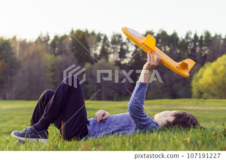 Calm teenager lying on meadow, holding airplane toy over body and looking away at forest, playing and dreaming. 107191227
