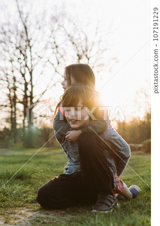 Little sister hugging brother from rear and looking away while teen sitting on lawn, preparing for carry girl on back. 107191229