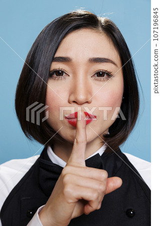 Serious asian woman in waitress uniform holding forefinger on lips while showing tsss gesture and looking at camera. Young receptionist asking for silence closeup portrait 107196845