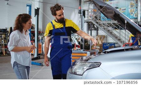 Dolly out shot of repairman assisting client with car maintenance in repair shop. Employee in garage workplace looking over automobile parts with woman, repairing her vehicle during yearly checkup Dolly out shot of repairman assisting client with car maintenance in repair shop. Employee in garage workplace looking over automobile parts with woman, repairing her vehicle during yearly checkup 107196856