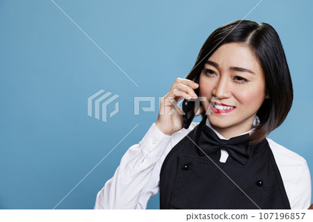 Hotel smiling cheerful asian woman receptionist wearing uniform speaking on smartphone. Joyful waitress answering call and chatting with catering service customer on mobile phone 107196857