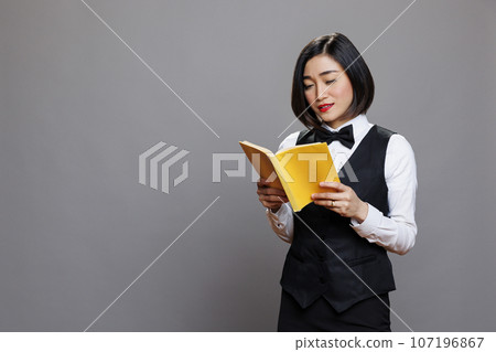 Thoughtful asian waitress dressed in professional uniform reading book with yellow cover. Young attractive woman receptionist holding softcover journal while posing in studio Thoughtful asian waitress dressed in professional uniform reading book with yellow cover. Young attractive woman receptionist holding softcover journal while posing in studio 107196867