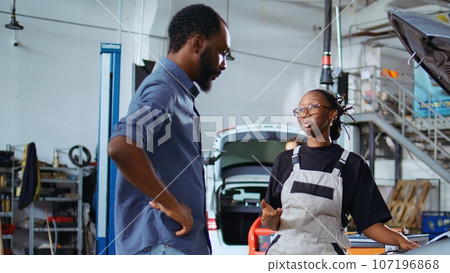 Mechanic in garage workspace fixing car for customer, thoroughly checking underneath vehicle hood, zoom out shot. Employee doing yearly maintenance on client automobile in auto repair shop Mechanic in garage workspace fixing car for customer, thoroughly checking underneath vehicle hood, zoom out shot. Employee doing yearly maintenance on client automobile in auto repair shop 107196868