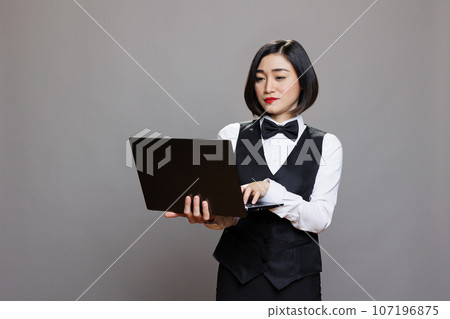 Young asian waitress using laptop, checking cafe online menu and managing catering service. Attractive restaurant receptionist wearing professional uniform and holding portable computer in studio 107196875