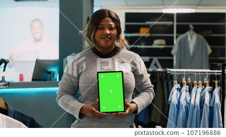 Young store manager showing tablet with green screen, advertising fashion boutique with chroma key display. Female employee using isolated mockup template with blank copyspace. 107196886