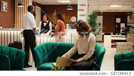 Busy hotel manager receiving cup of coffee from employee while checking paperwork of guests waiting to be checked in. Receptionist offers room access to couple after booking process is finalized 107196913