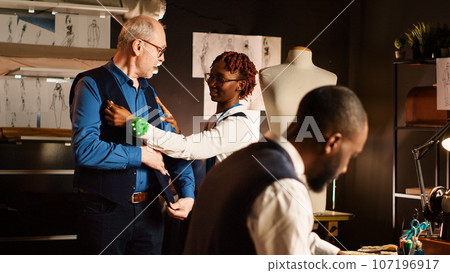 Old customer trying on suit jacket in workshop, team of couturiers ensuring fit size before designing tailored clothes. Client checking clothing item measurement, small business. Handheld shot. Old customer trying on suit jacket in workshop, team of couturiers ensuring fit size before designing tailored clothes. Client checking clothing item measurement, small business. Handheld shot. 107196917