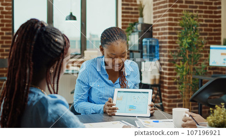 Female manager showing research plan, doing presentation in boardroom coworking space with tablet. Group of colleagues discussing new strategy with business results for development. 107196921