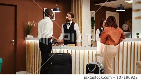 Professional friendly receptionist team greeting tourists on holiday trip in resort lobby. Helpful concierge personnel welcoming guests at hotel reception front desk during check in process 107196922