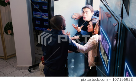Impatient shoppers fighting staff to enter clothing store, pushing african american security guard on black friday. Agent holding back angry eager crowd, trying to keep door closed. Crowd control. Impatient shoppers fighting staff to enter clothing store, pushing african american security guard on black friday. Agent holding back angry eager crowd, trying to keep door closed. Crowd control. 107196938