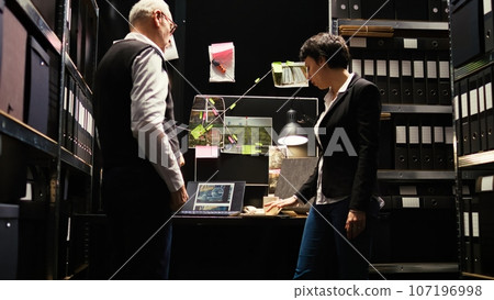 Law agents review police archive records in incident room, gathering intelligence from witness statements. Detectives team evaluating evidence authenticity, inspecting forensic report. 107196998