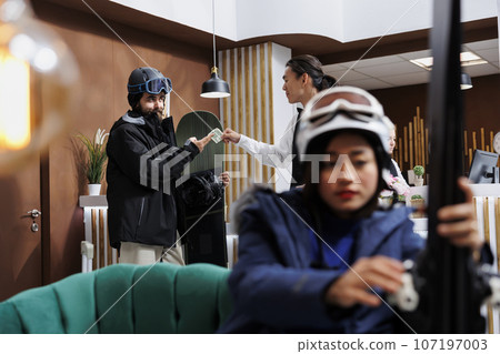 Image shows woman adjusting skiing equipment on sofa while man with snowboard gives money to concierge for good customer service. Male tourist tipping staff in hotel lobby as lady waits on couch. Image shows woman adjusting skiing equipment on sofa while man with snowboard gives money to concierge for good customer service. Male tourist tipping staff in hotel lobby as lady waits on couch. 107197003